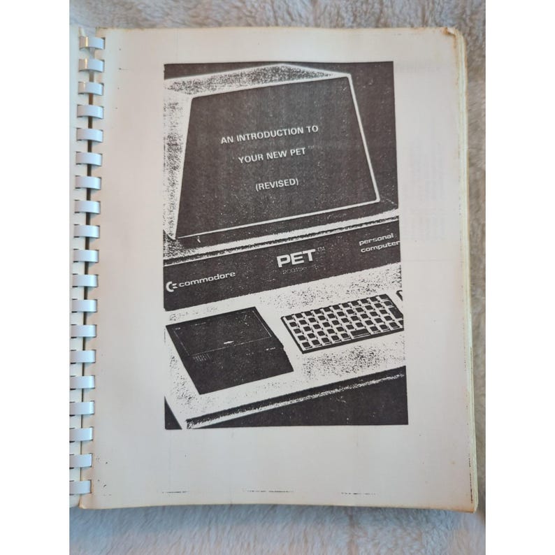 Pu&ograve; includere: Un libro aperto con rilegatura a spirale, con un'immagine in bianco e nero di un computer personale Commodore PET vintage. Lo schermo mostra il testo "AN INTRODUCTION TO YOUR NEW PET (REVISED)". Sono visibili anche la tastiera e un'unit&agrave; a cassette.