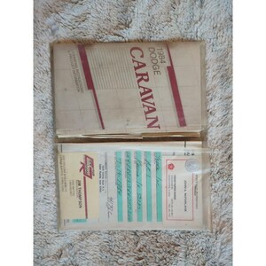 May include: A stack of paper manuals for a 1984 Dodge Caravan. The top manual has the title "1984 Dodge Caravan" printed in red and white. The bottom manual has a sticker with the text "Jim Thompson" and "Cedar Rapids Dodge" printed on it.