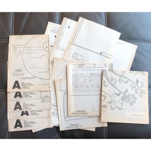 May include: A stack of vintage American Pass-Times magazines with cross-stitch patterns, including a floral design and a pattern with the text "Elizabeth". The magazines are on a brown surface.