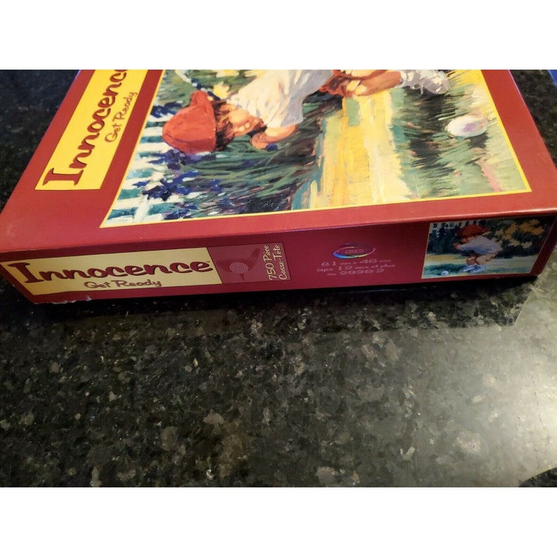 May include: A red box with the words "Innocence Get Ready" on the front. The box is for a 750 piece jigsaw puzzle with a picture of a child in a red hat playing baseball. The box also has the words "750 Piece  Cross-Tile" and "61 x 46 cm  24 x 18 ins" printed on it.