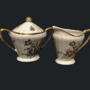 May include: A white sugar bowl and creamer set with a gold trim and black floral design. The set is made of porcelain and has a vintage look.