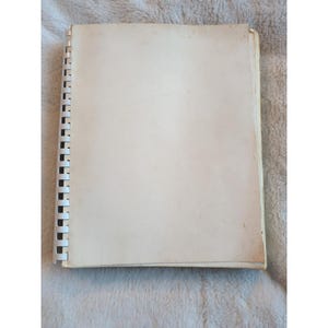 Pu&ograve; includere: Un quaderno a spirale aperto, con pagine bianco sporco. Il quaderno &egrave; aperto, rivelando pagine vuote. La rilegatura &egrave; nera e le pagine sono leggermente ingiallite. Il quaderno &egrave; appoggiato su una superficie strutturata.