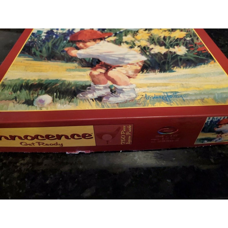 May include: A 750 piece jigsaw puzzle box with a red background and the words "Innocence Get Ready" in white. The box features a colorful illustration of a young child playing golf in a green grassy field with flowers.