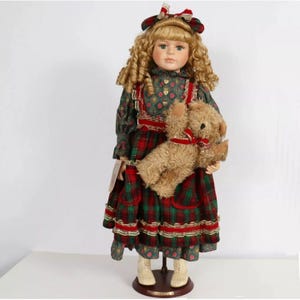 May include: A porcelain doll with blonde curly hair wearing a green and red tartan dress with a red ribbon and a teddy bear. The doll is standing on a wooden base.
