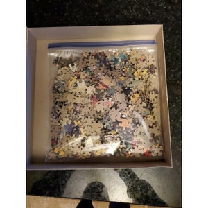 May include: A plastic bag filled with a jigsaw puzzle pieces. The pieces are various colors and shapes. The bag is in a cardboard box.