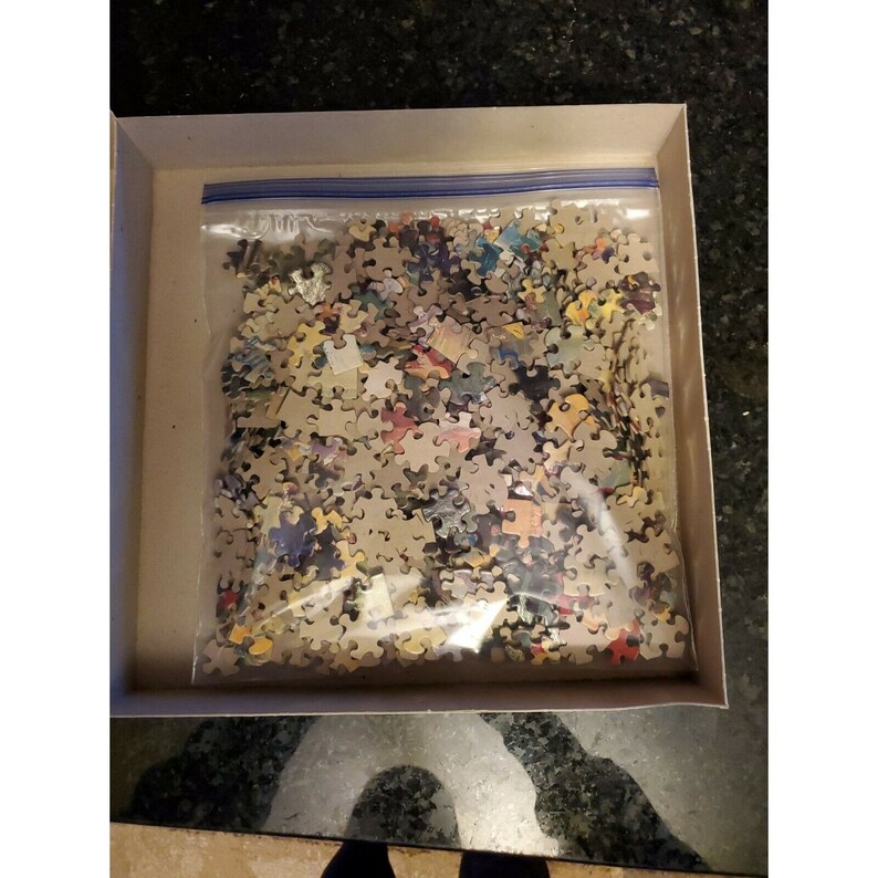 May include: A plastic bag filled with a jigsaw puzzle pieces. The pieces are various colors and shapes. The bag is in a cardboard box.