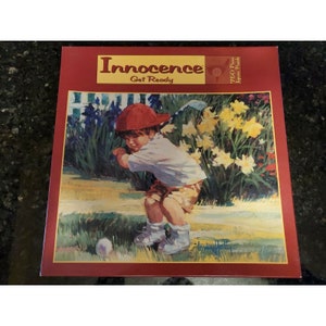 May include: A 750 piece jigsaw puzzle box featuring a painting of a young boy in a red hat and white shirt swinging a golf club. The boy is in a garden setting with yellow flowers and green grass. The box has the title "Innocence Get Ready" printed on the front.