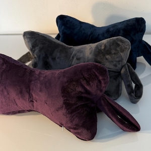 May include: Three plush neck pillows in shades of purple, gray, and navy blue. Each pillow has a unique shape with extended sides. The pillows are made of a soft, velvety material.