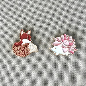 May include: Two enamel pins featuring a red fox with a white tail and a white hedgehog with a pink flower on its back.