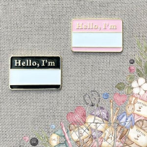 May include: Two enamel pins with a name tag design. The pins say "Hello, I'm" with a blank space for a name. One pin is black with a white background and the other is pink with a white background.