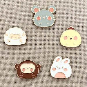May include: Five enamel pins featuring cartoon animals. The pins are in the shape of a mouse, a sheep, a chick, a bunny, and a monkey. The pins are all gold-colored with white, gray, yellow, pink, and brown accents.