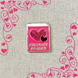 May include: A pink enamel pin in the shape of a book with the words "ROMANCE READER" in gold lettering. The book cover features a large glittery heart and a smaller black heart.