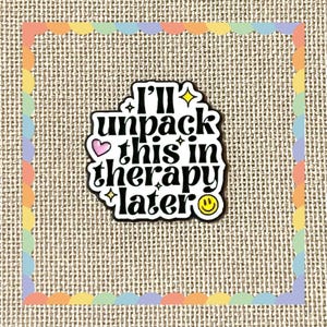 May include: A white enamel pin with a black border featuring the black text I'll unpack this in therapy later. The design includes a small pink heart, yellow four-pointed stars, and a yellow smiley face icon at the bottom right corner.