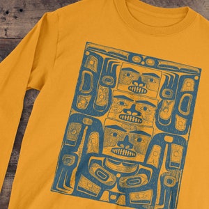 May include: Mustard yellow long-sleeve shirt featuring a blue graphic print of a stylized totem design. The artwork depicts three faces stacked vertically, with intricate details and patterns. The shirt is made of a soft material.