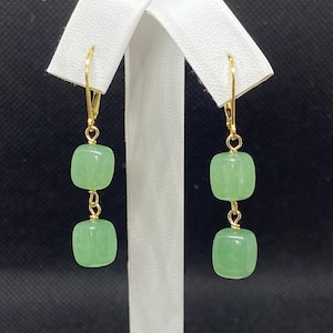 May include: A pair of gold-colored leverback earrings with two light green, square-shaped beads. The beads are connected by gold-colored wire. The earrings are displayed against a white background.