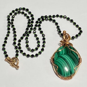 May include: A necklace featuring a large, oval-shaped green malachite pendant encased in gold-toned wire wrapping. The chain consists of small, faceted black beads strung on a bright green cord, finished with a gold-toned toggle clasp closure.
