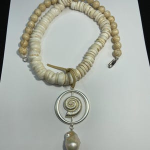 May include: A cream-colored necklace featuring a pendant with a spiral design and a large, pear-shaped pearl. The necklace is made of white shell discs and beige beads, with a silver clasp and a leather cord.