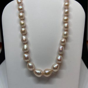 May include: A necklace of lustrous, off-white pearls with a subtle pink hue. The pearls are irregularly shaped, strung together with a delicate pink string, and displayed on a white jewelry stand. The necklace is a classic piece of jewelry.