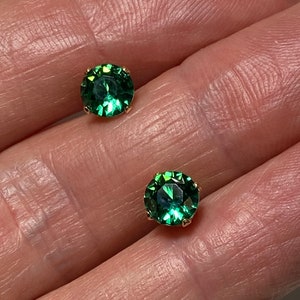 May include: A pair of gold-colored stud earrings, each featuring a round, faceted, emerald-green gemstone. The earrings are set in a classic prong setting, showcasing the vibrant green color of the stones.