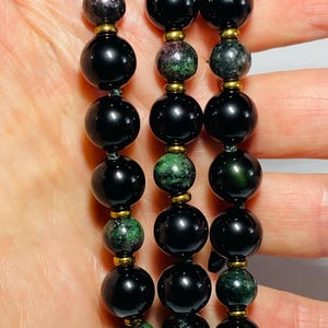 May include: Close-up of three strands of beaded jewelry. The beads alternate between large, glossy black spheres and smaller, green and purple speckled beads. Gold-colored spacers separate the beads. The jewelry is held in a person's hand.