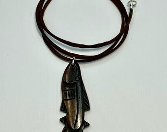 May include: A brown wooden fish pendant necklace with a dark brown suede cord. The fish pendant has carved details and a silver-colored bail. The necklace is displayed against a white background.