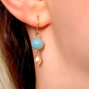 Op de afbeelding: Een paar oorhangers met een lichtblauwe edelsteen en een witte parel. De oorhangers zijn gemaakt van goudkleurig metaal.