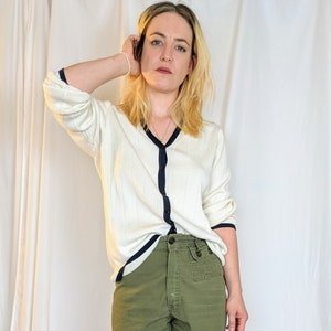 May include: A woman wearing a white cardigan with a navy blue trim and green cargo shorts. She is wearing beige lace-up shoes.