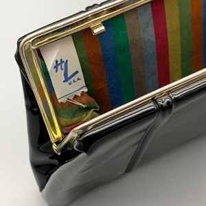 Puede incluir: Clutch de charol negro con marco dorado y forro de tela a rayas. El forro tiene una etiqueta que dice "H.L. U.S.A."