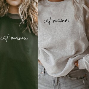 Pu&ograve; includere: Due donne indossano felpe con il testo "cat mama" in corsivo bianco. Una felpa &egrave; verde scuro e l'altra &egrave; grigio chiaro.