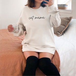 Pu&ograve; includere: Una felpa bianca oversize con il testo "cat mama" in corsivo nero.