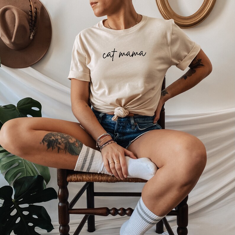Pu&ograve; includere: Una donna indossa una maglietta beige chiaro con il testo "cat mama" in caratteri neri. &Egrave; seduta su una sedia di legno con le gambe incrociate e indossa calzini bianchi con strisce nere.