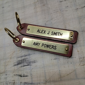 May include: Two personalized leather keychains with brass nameplates. One reads "ALEX J SMITH" and the other "AMY POWERS". The keychains are brown with gold-colored key rings.