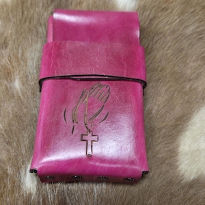 May include: A pink leather wallet with a praying hands and cross design. The wallet is closed with a black elastic band. The background is a brown and white fur-like material.