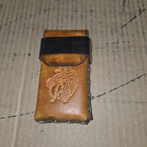 Könnte beinhalten: Eine braune Ledertasche mit einem schwarzen Band und einem geprägten Drachen auf der Vorderseite. Die Tasche ist mit Metallnieten befestigt und scheint handgefertigt zu sein. Das Leder hat einen abgenutzten, Vintage-Look.