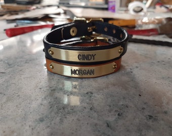 Puede incluir: Dos collares de perro de cuero con placas de nombre de latón. El collar superior es negro con el nombre "CINDY" grabado en la placa. El collar inferior es marrón con el nombre "MORGAN" grabado en la placa.
