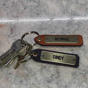 Puede incluir: Dos llaveros de cuero con etiquetas de nombre de metal. El llavero superior es marrón con el nombre "GEORGE" en la etiqueta. El llavero inferior es negro con el nombre "CINDY" en la etiqueta.