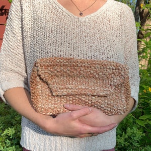 May include: A woven clutch purse with a brown and beige color scheme. The clutch has a flap closure and a textured surface.