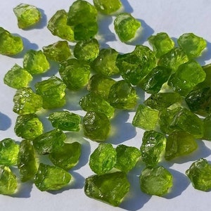 Pode incluir: Uma coleção de pequenas pedras preciosas de peridoto em bruto, em vários tons de verde. As pedras preciosas estão espalhadas sobre uma superfície branca, com sombras criadas pela luz natural.