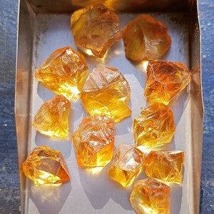 May include: A collection of 12 rough, faceted, amber-colored gemstones in a metal tray. The gemstones are various sizes and shapes, with some having a more rounded shape and others having a more angular shape.