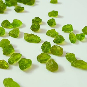 May include: A collection of small, rough, green peridot gemstones. The stones are scattered on a white surface.