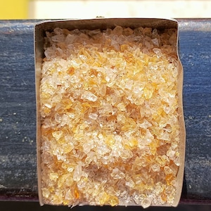 Puede incluir: Primer plano de un pequeño recipiente metálico lleno de cristales translúcidos de color ámbar, de forma irregular.