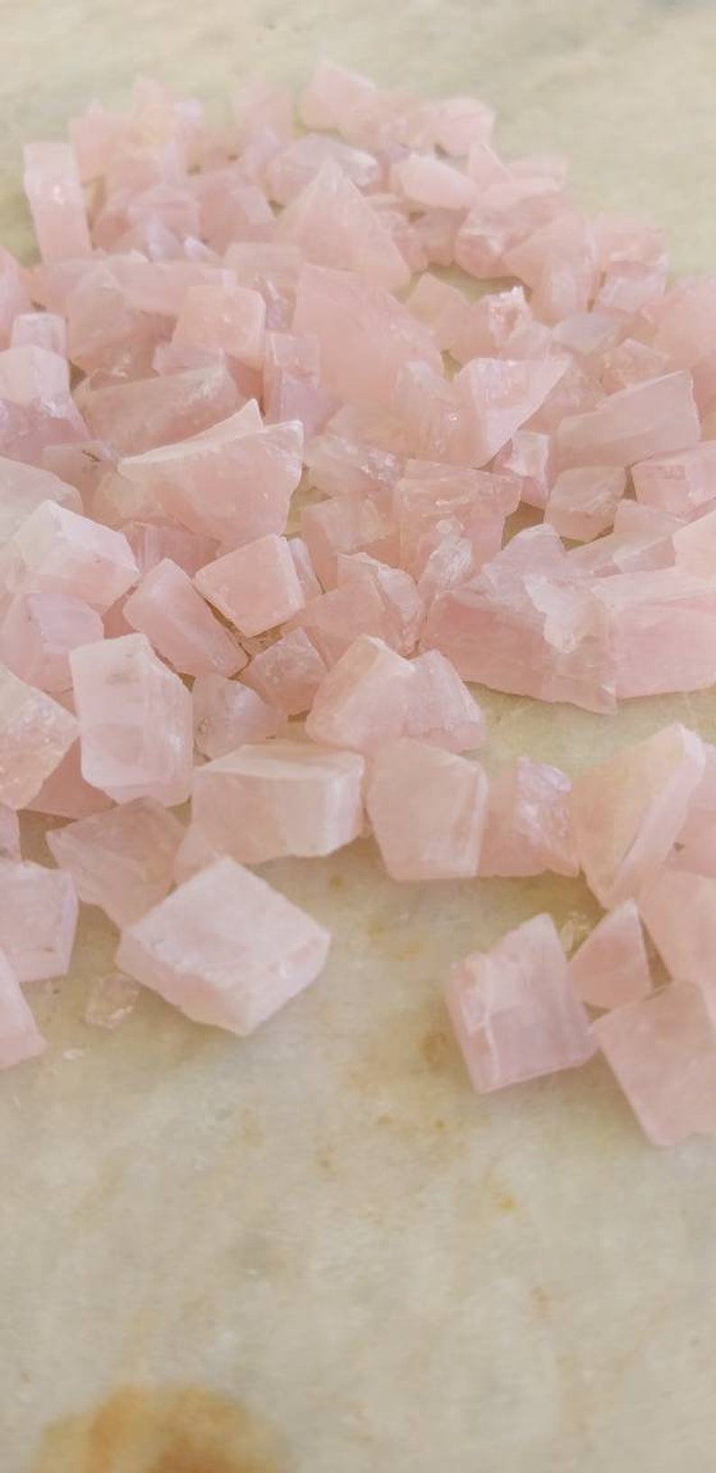 May include: A large pile of pink rose quartz crystals, tumbled and polished, with irregular shapes and sizes. The crystals are scattered on a light beige surface.