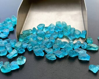 May include: A scoop of loose, faceted, teal blue gemstones. The gemstones are scattered on a dark gray surface.