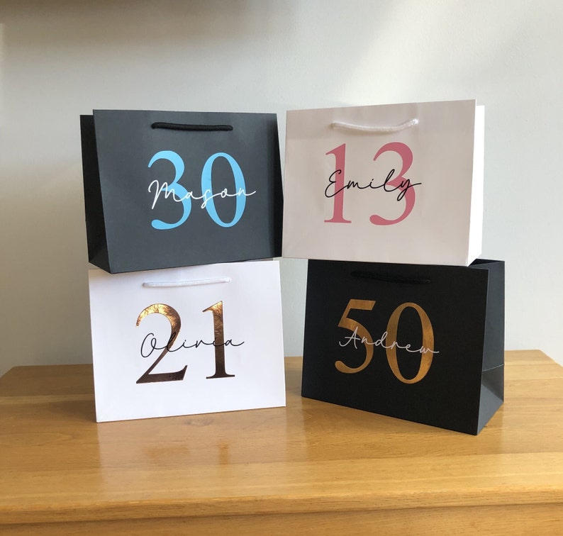 Puede incluir: Cuatro bolsas de regalo en blanco y negro, cada una con una edad y un nombre diferentes. Las bolsas tienen asas negras o blancas. Las edades son 30, 13, 21 y 50. Los nombres son Mason, Emily, Olivia y Andrew.