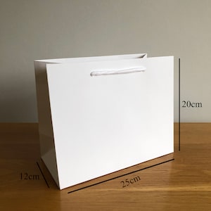 Puede incluir: Una bolsa de regalo blanca con acabado brillante. La bolsa es rectangular, mide 25 cm de largo, 20 cm de alto y 12 cm de ancho. Tiene un asa de cuerda blanca. La bolsa est&aacute; sobre una superficie de madera.