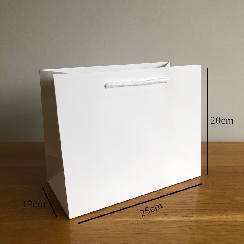 Puede incluir: Una bolsa de regalo blanca con acabado brillante. La bolsa es rectangular, mide 25 cm de largo, 20 cm de alto y 12 cm de ancho. Tiene un asa de cuerda blanca. La bolsa est&aacute; sobre una superficie de madera.
