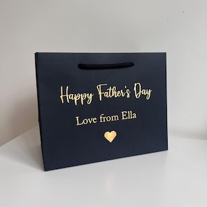 May include: A navy blue gift bag with a black rope handle. The bag features gold lettering that reads "Happy Father's Day" and "Love from Ella", with a gold heart below. The bag is on a white surface.