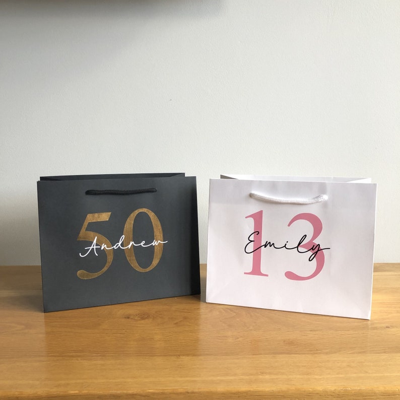 Puede incluir: Dos bolsas de regalo, una gris oscuro y otra blanca, est&aacute;n una al lado de la otra sobre una superficie de madera. La bolsa gris oscuro tiene el n&uacute;mero "50" en dorado y el nombre "Andrew" en escritura blanca. La bolsa blanca tiene el n&uacute;mero "13" en rosa y el nombre "Emily" en escritura negra.