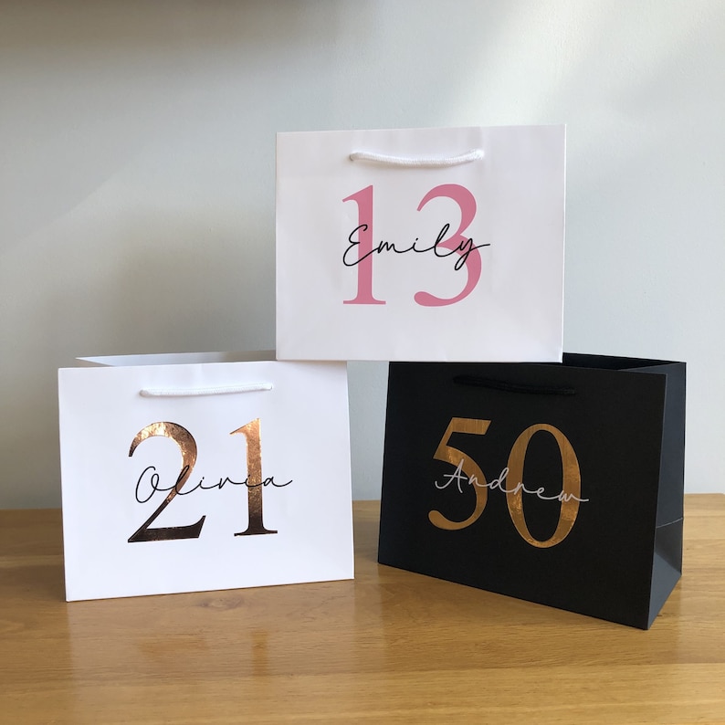 Puede incluir: Tres bolsas de regalo con asas de cuerda blanca. Una bolsa negra con texto dorado que dice "50 Andrew". Dos bolsas blancas con texto rosa y dorado. Una dice "13 Emily", la otra "21 Olivia".
