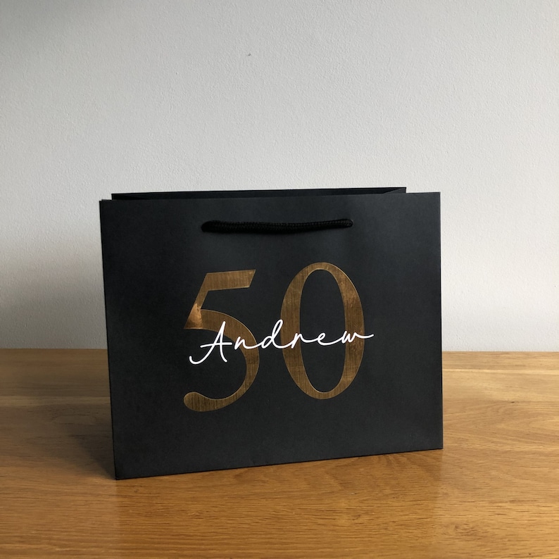Puede incluir: Bolsa de regalo negra con el n&uacute;mero '50' dorado y el nombre 'Andrew' en escritura blanca. La bolsa tiene un asa de cord&oacute;n negro. Ideal para un 50 cumplea&ntilde;os. Sobre una superficie de madera.